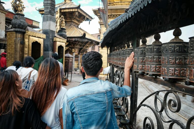 People touching prayer wheels at a temple, with intricate carvings and Nepali script, set against a backdrop of traditional architecture.