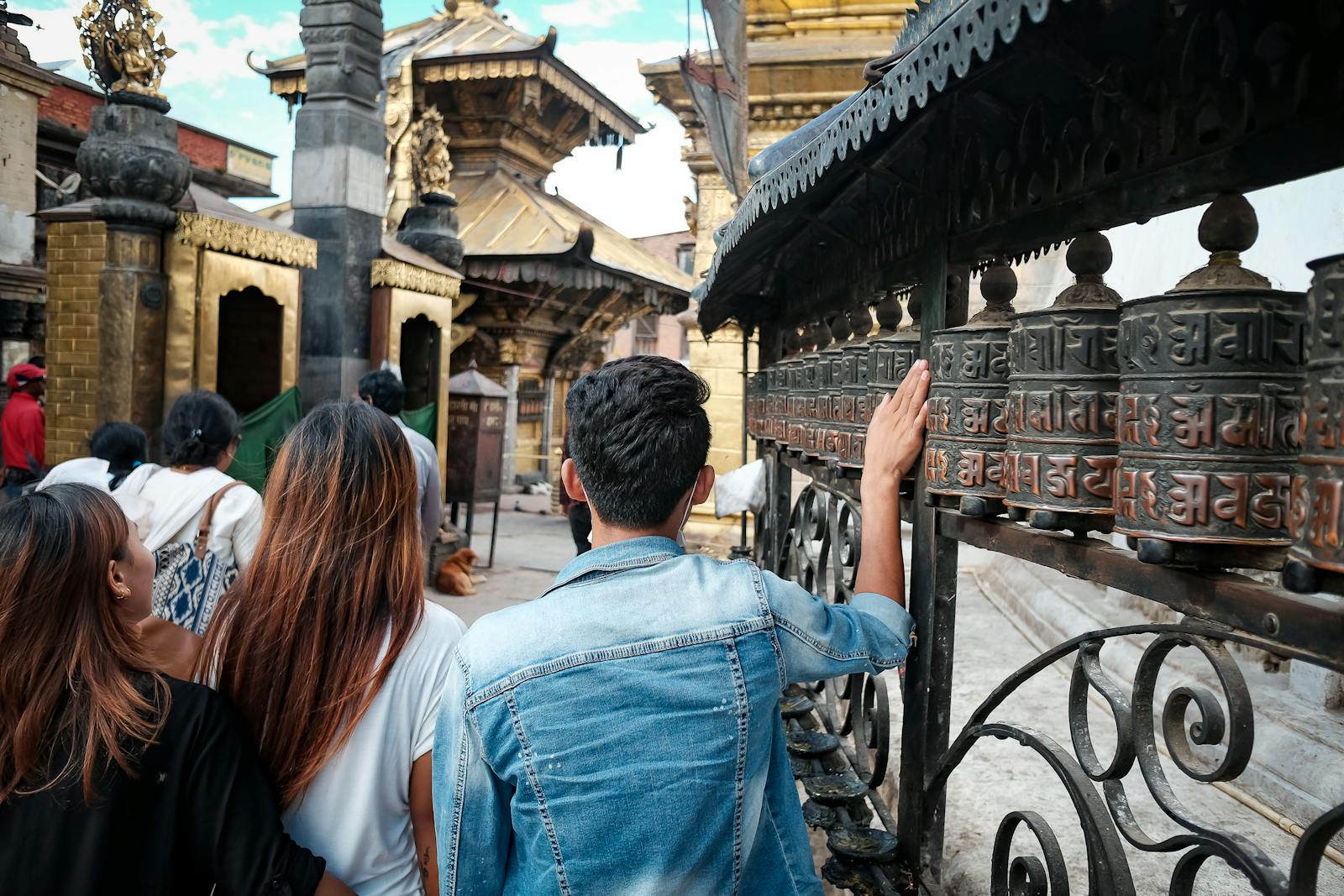 People touching prayer wheels at a temple, with intricate carvings and Nepali script, set against a backdrop of traditional architecture.