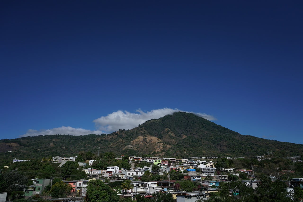 A panoramic view of a small town with colorful buildings at the foot of a lush, green mountain under a clear blue sky with minimal clouds.