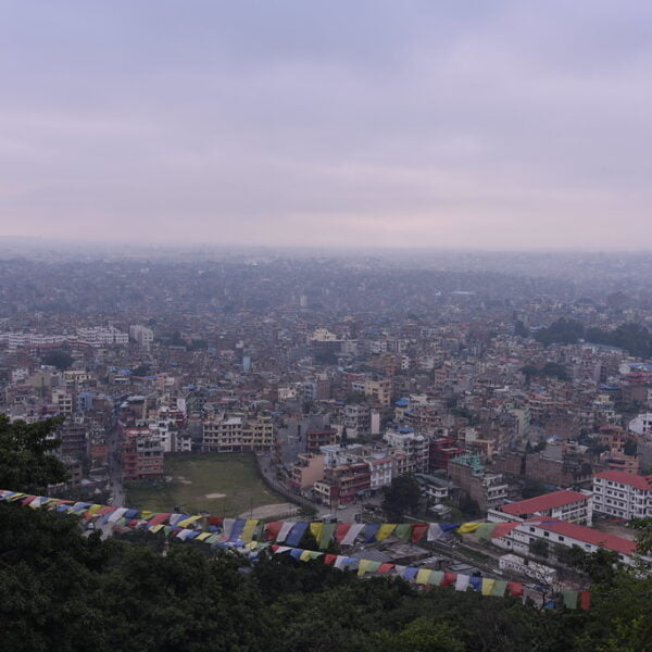 A panoramic view of a densely populated city with buildings extending into the horizon, viewed from a hilltop with prayer flags in the foreground.