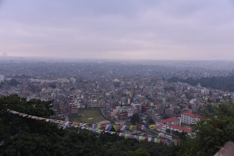 A panoramic view of a densely populated city with buildings extending into the horizon, viewed from a hilltop with prayer flags in the foreground.