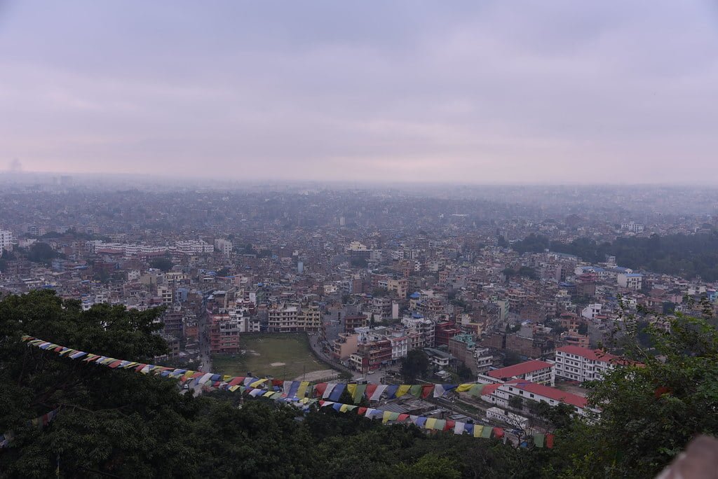 A panoramic view of a densely populated city with buildings extending into the horizon, viewed from a hilltop with prayer flags in the foreground.