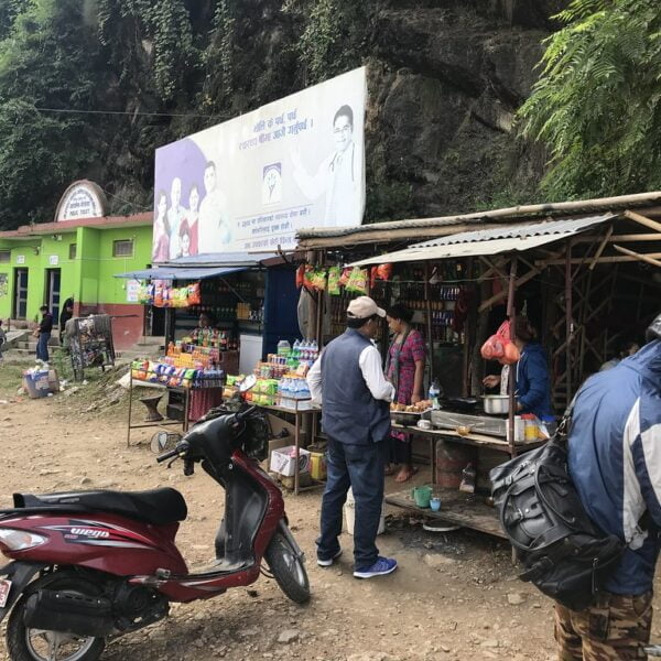 A bustling roadside market in a mountainous area with people engaging in daily commerce. There's a man wearing a cap and jacket observing a stall with various snacks and a motorcyclist with a helmet, beside a parked red scooter. The background features green-painted buildings and lush vegetation with a large banner overhead.