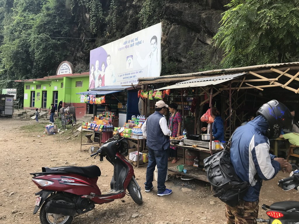 A bustling roadside market in a mountainous area with people engaging in daily commerce. There's a man wearing a cap and jacket observing a stall with various snacks and a motorcyclist with a helmet, beside a parked red scooter. The background features green-painted buildings and lush vegetation with a large banner overhead.