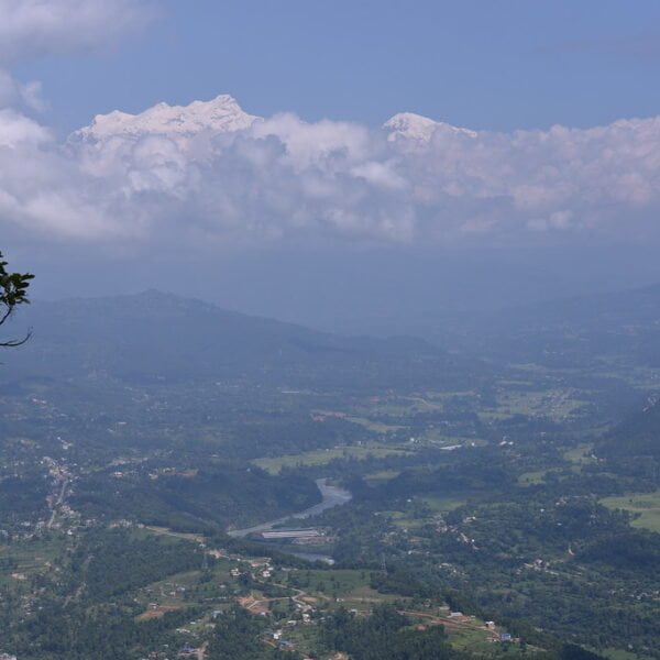 A panoramic view of a valley with a river, towns, and greenery, overlooked by a snow-capped mountain partially shrouded in clouds, and foliage in the foreground.