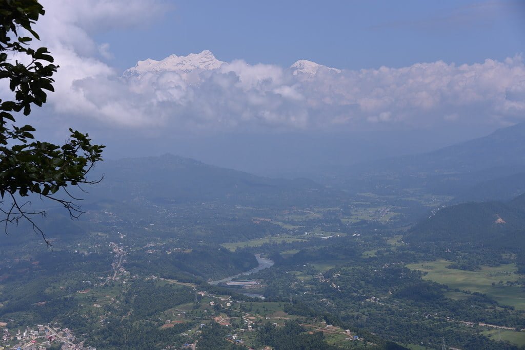 A panoramic view of a valley with a river, towns, and greenery, overlooked by a snow-capped mountain partially shrouded in clouds, and foliage in the foreground.