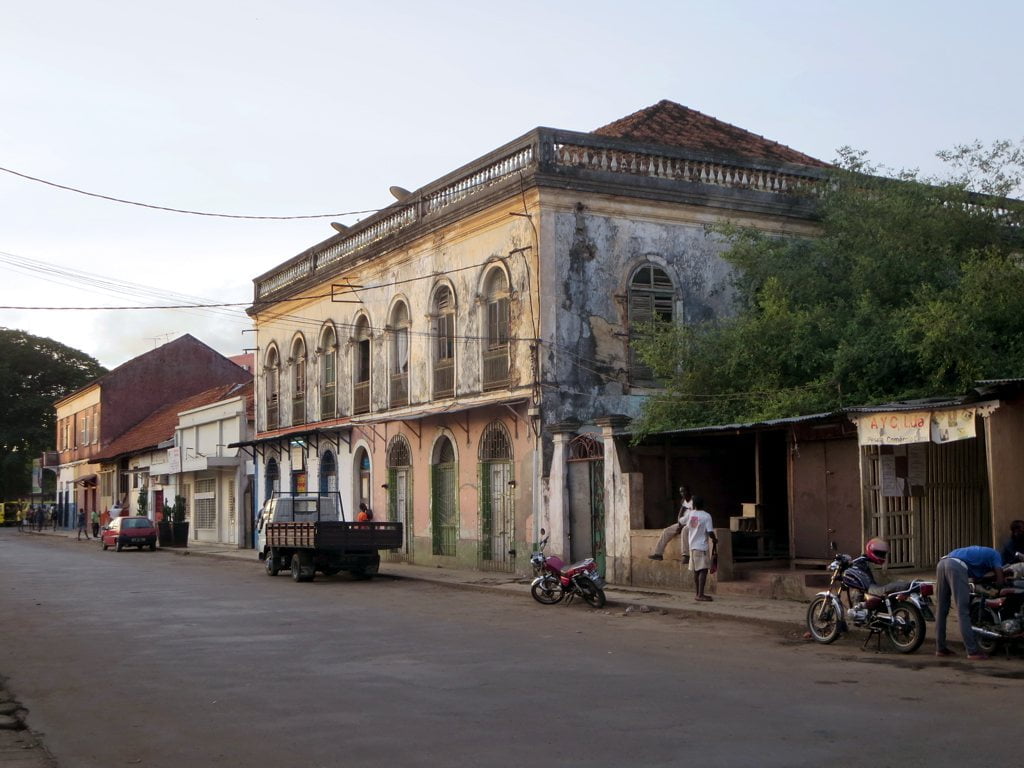 A weathered two-story colonial building with arched windows on a street corner. The building shows signs of neglect with peeling paint and a partially overgrown facade. A few people walk by and motorcycles are parked along the curb. Shops with closed shutters are visible on the ground floor, and the sky is clear at dusk.