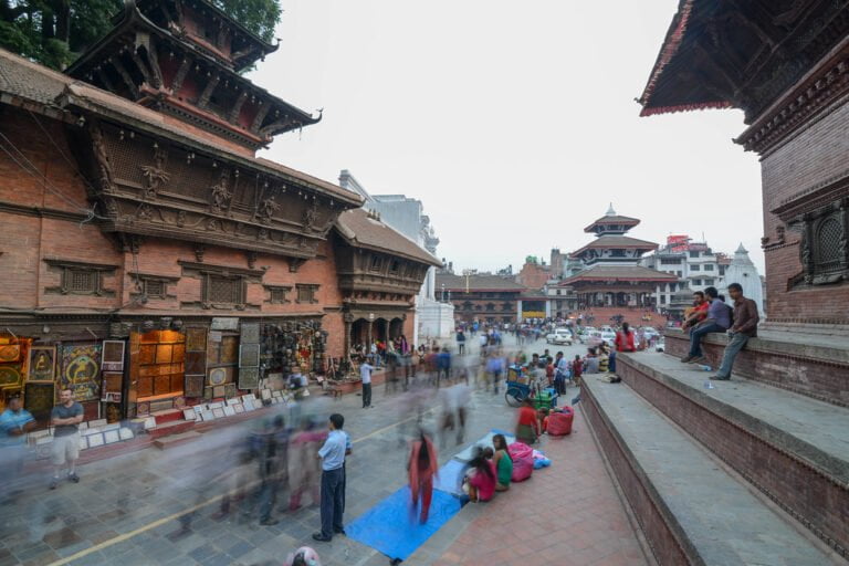 A bustling market scene in a Nepalese square with traditional architecture, people sitting on steps, and motion blur suggesting movement.