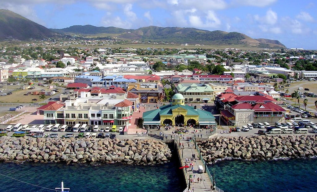 Aerial view of a coastal town with a pier extending into the sea, colorful buildings, and a backdrop of green hills under a blue sky.