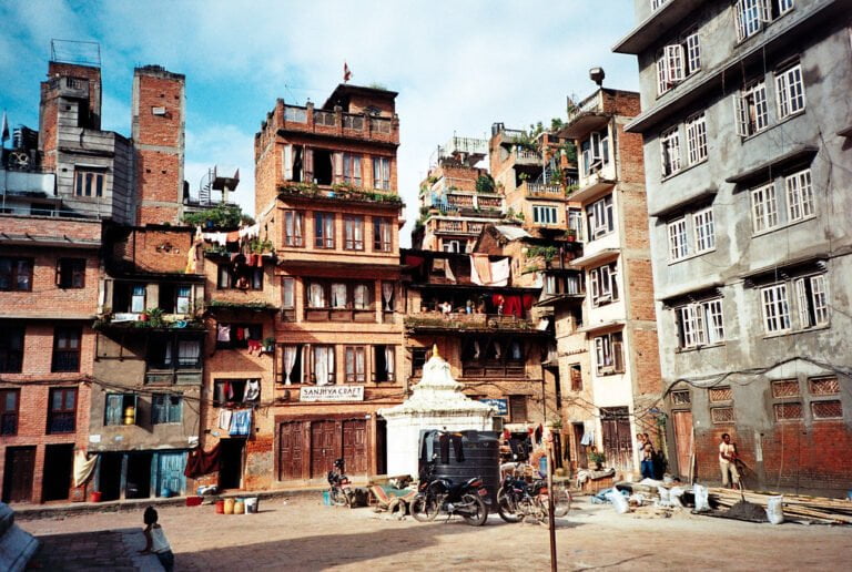 A vibrant street scene with multi-story buildings closely packed in an urban area, displaying various architectural styles and balconies adorned with plants. People are seen going about their daily activities with bicycles and a rickshaw present on the street.