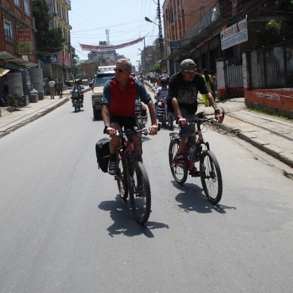 Two people cycling on a street in an urban area with shops and pedestrians visible on the sidelines.