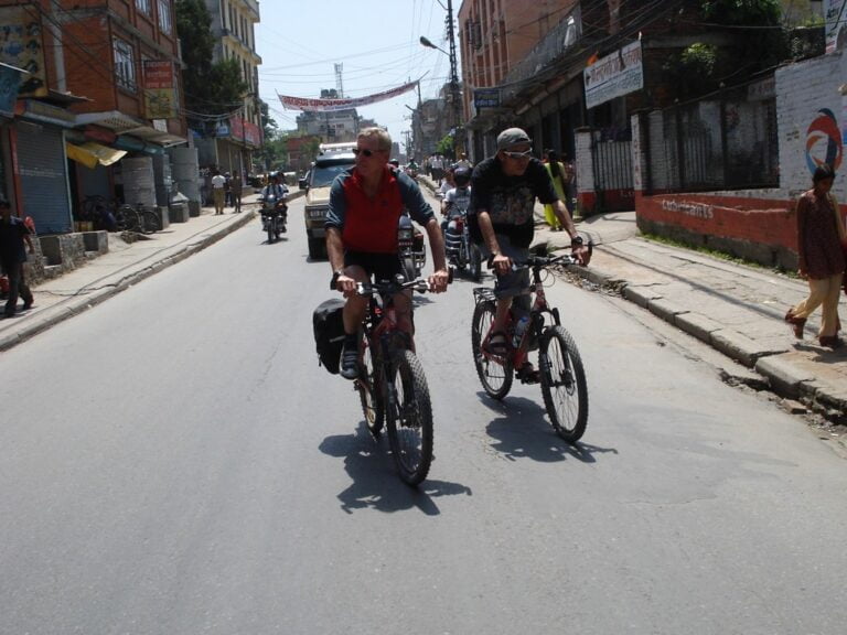 Two people cycling on a street in an urban area with shops and pedestrians visible on the sidelines.
