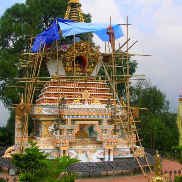 A colorful Buddhist stupa under renovation with scaffolding and blue tarpaulin around it, set against a backdrop of greenery and overcast skies.