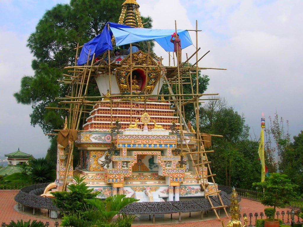 A colorful Buddhist stupa under renovation with scaffolding and blue tarpaulin around it, set against a backdrop of greenery and overcast skies.