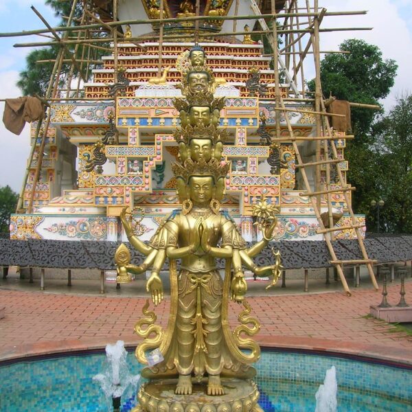 Alt text: A golden multi-armed statue stands in front of a partially constructed colorful Buddhist stupa, with scaffolding visible. The statue is adjacent to a small blue-tiled fountain and the scene is surrounded by greenery.
