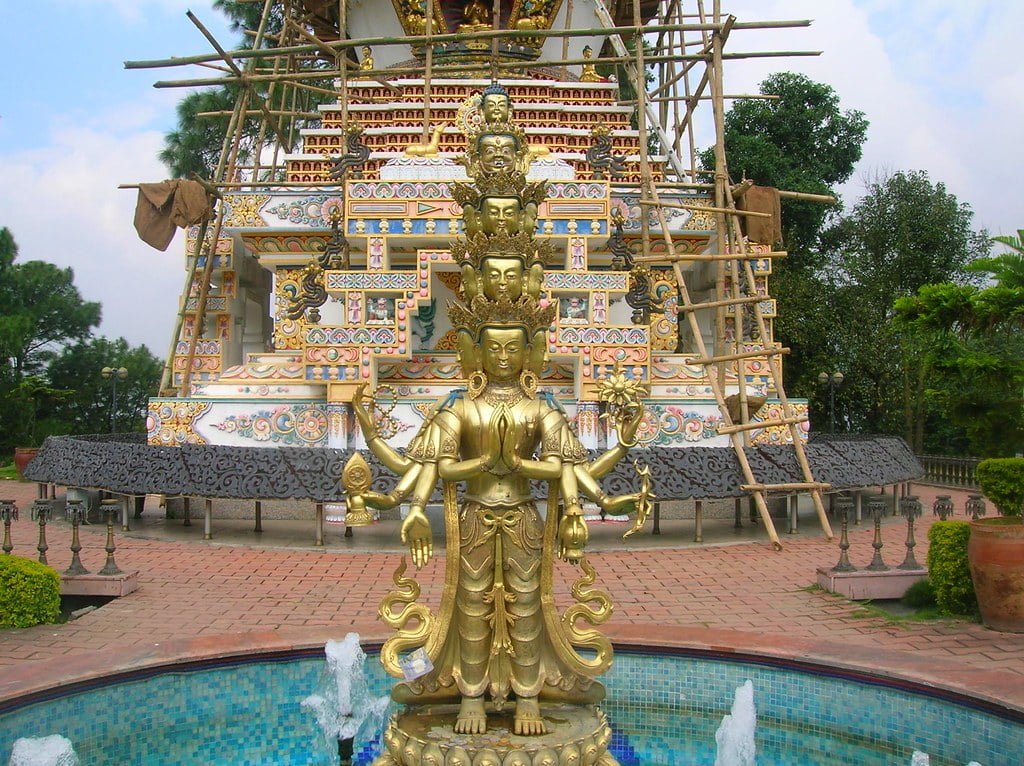 Alt text: A golden multi-armed statue stands in front of a partially constructed colorful Buddhist stupa, with scaffolding visible. The statue is adjacent to a small blue-tiled fountain and the scene is surrounded by greenery.