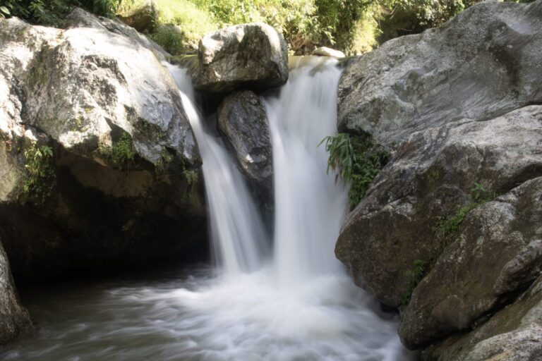A small waterfall cascading between large rocks with a smooth, silky appearance of the water created by a long exposure photography technique, surrounded by green vegetation.