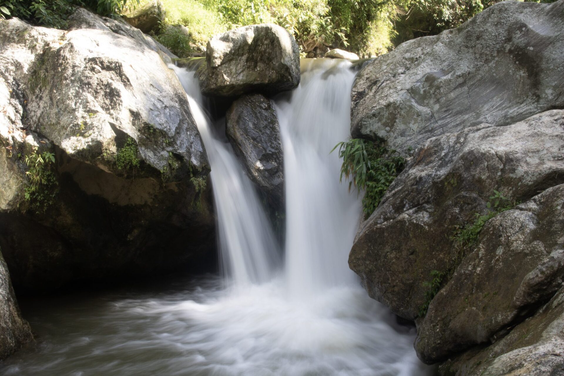 A small waterfall cascading between large rocks with a smooth, silky appearance of the water created by a long exposure photography technique, surrounded by green vegetation.