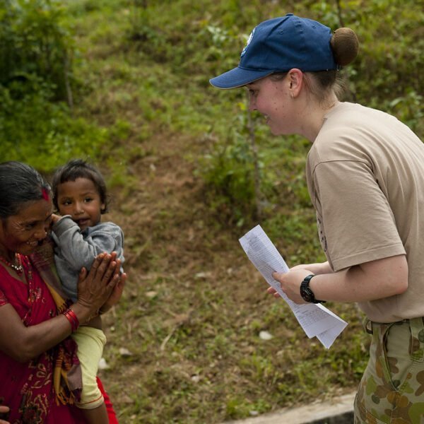 A woman in a red traditional dress holding a child interacts with another woman in a baseball cap and beige shirt who is holding a piece of paper, in an outdoor setting with greenery in the background.