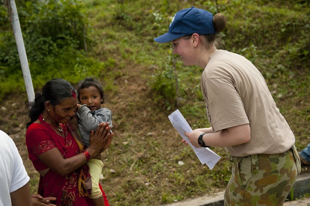 A woman in a red traditional dress holding a child interacts with another woman in a baseball cap and beige shirt who is holding a piece of paper, in an outdoor setting with greenery in the background.