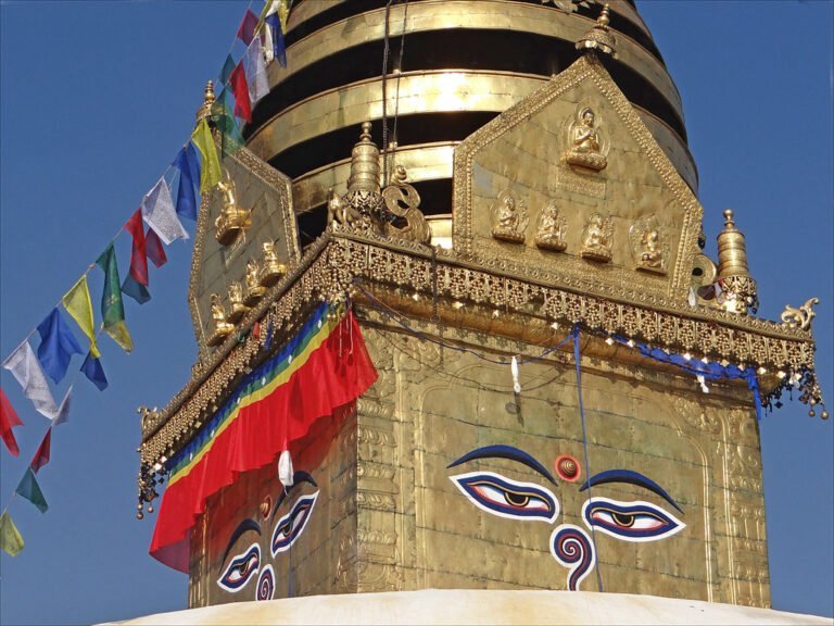 Alt text: A close-up of a golden stupa adorned with the iconic "Wisdom Eyes," colorful prayer flags, and intricate metalwork under a clear blue sky.