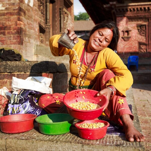 A smiling woman in traditional attire sits on the ground while sorting grain into colorful bowls at an outdoor market.