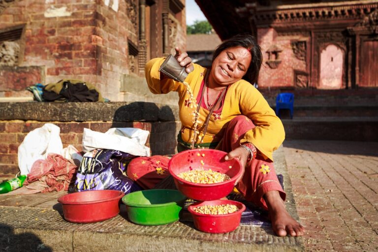 A smiling woman in traditional attire sits on the ground while sorting grain into colorful bowls at an outdoor market.