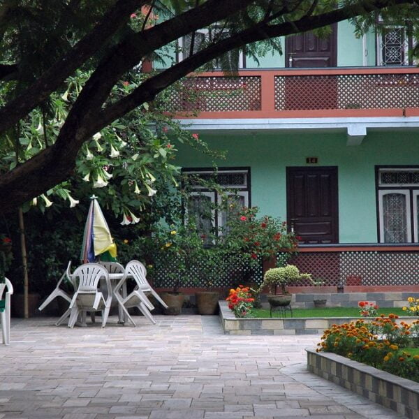 A tranquil garden setting with a paved walkway, a large shade tree, white plastic chairs around a closed umbrella, a green house with decorative windows, and colorful flowerbeds.
