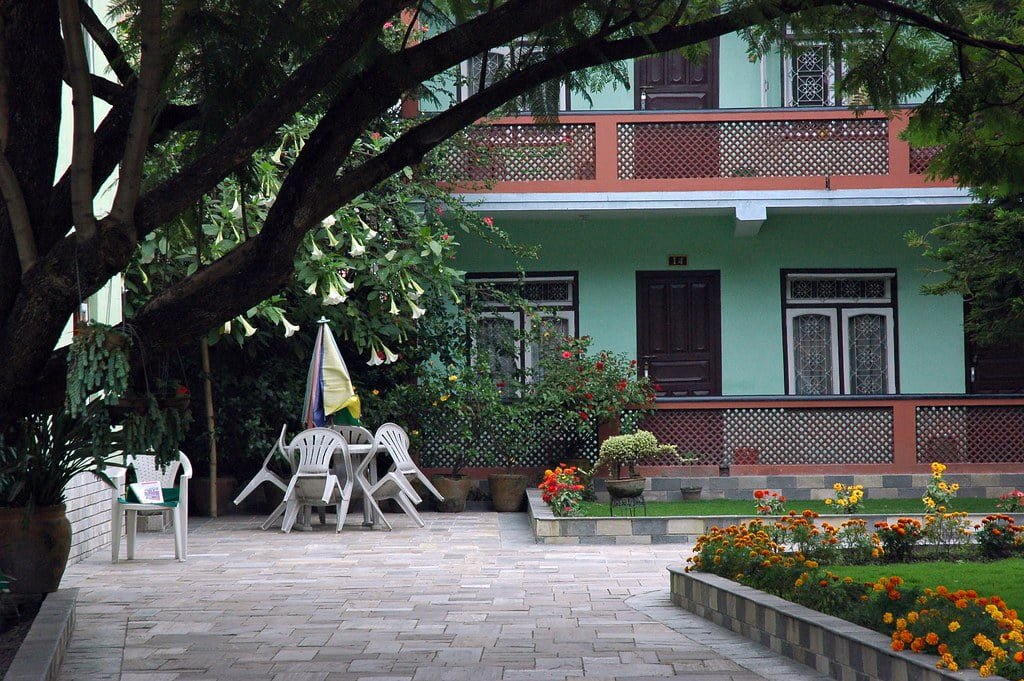 A tranquil garden setting with a paved walkway, a large shade tree, white plastic chairs around a closed umbrella, a green house with decorative windows, and colorful flowerbeds.