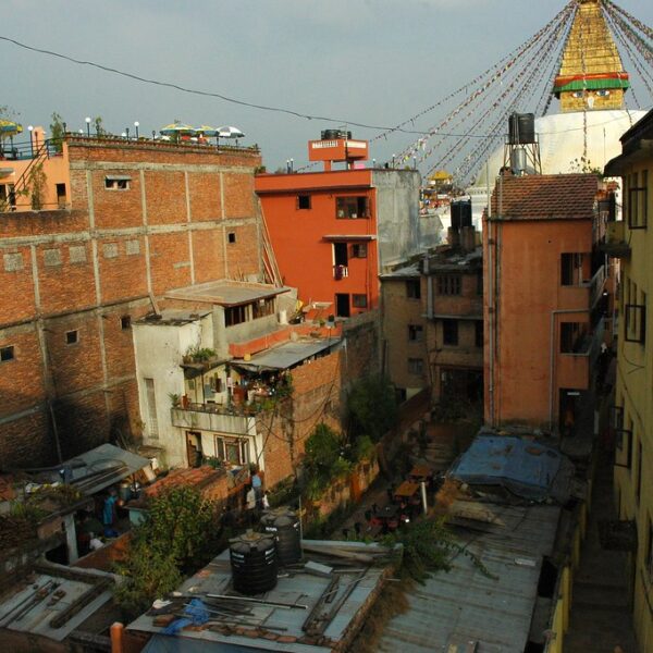 A cityscape showing tightly packed, multistory buildings with mixed brick and plaster exteriors, flat roofs adorned with water tanks, and a large Buddhist stupa visible in the background amid prayer flags.