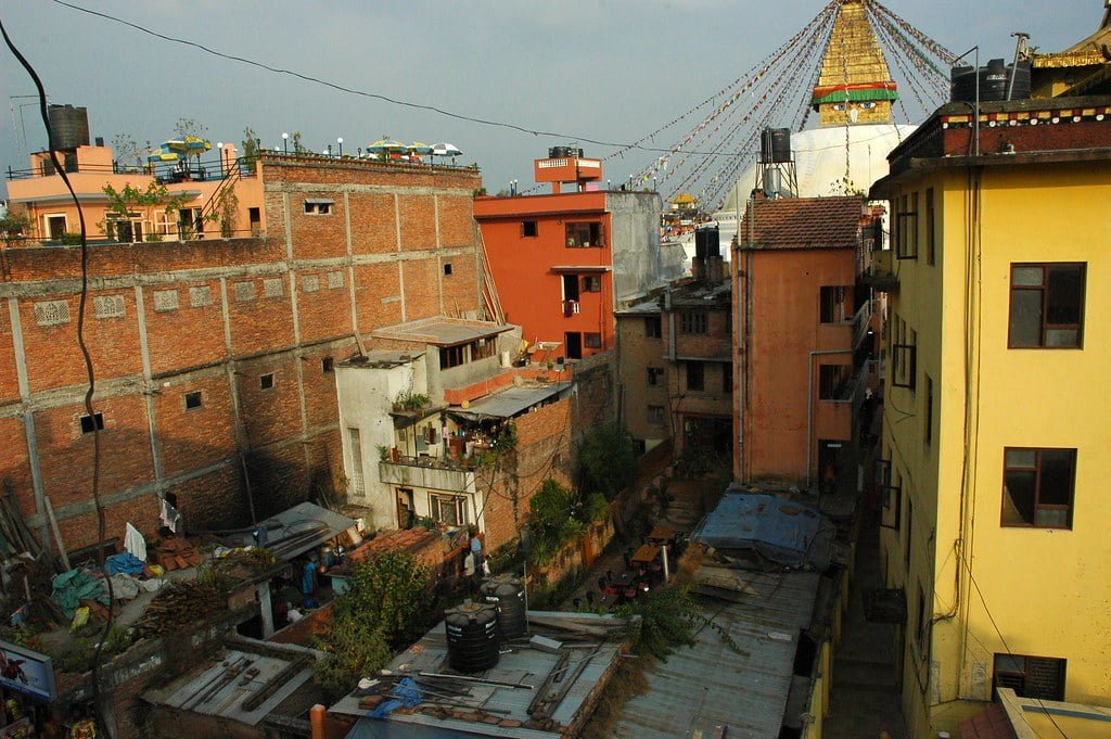 A cityscape showing tightly packed, multistory buildings with mixed brick and plaster exteriors, flat roofs adorned with water tanks, and a large Buddhist stupa visible in the background amid prayer flags.