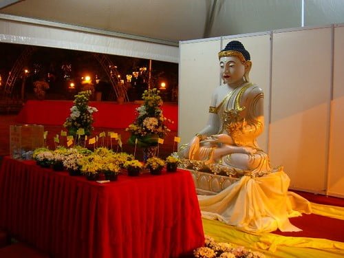 A statue of a seated Buddha in a meditative pose, adorned in golden attire, placed on a red-clothed table amidst floral arrangements inside a tented area.
