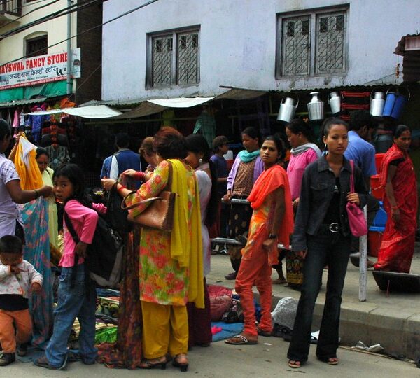 Street scene with people shopping at market stalls, some wearing traditional South Asian attire, with buildings and signs in the background.