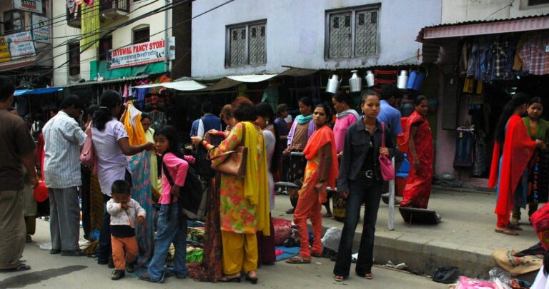 Street scene with people shopping at market stalls, some wearing traditional South Asian attire, with buildings and signs in the background.