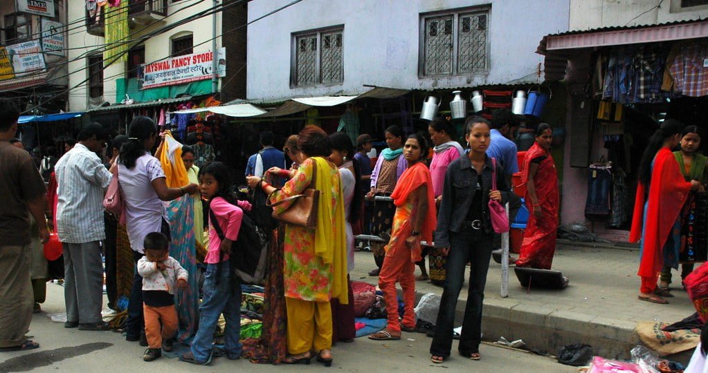 Street scene with people shopping at market stalls, some wearing traditional South Asian attire, with buildings and signs in the background.