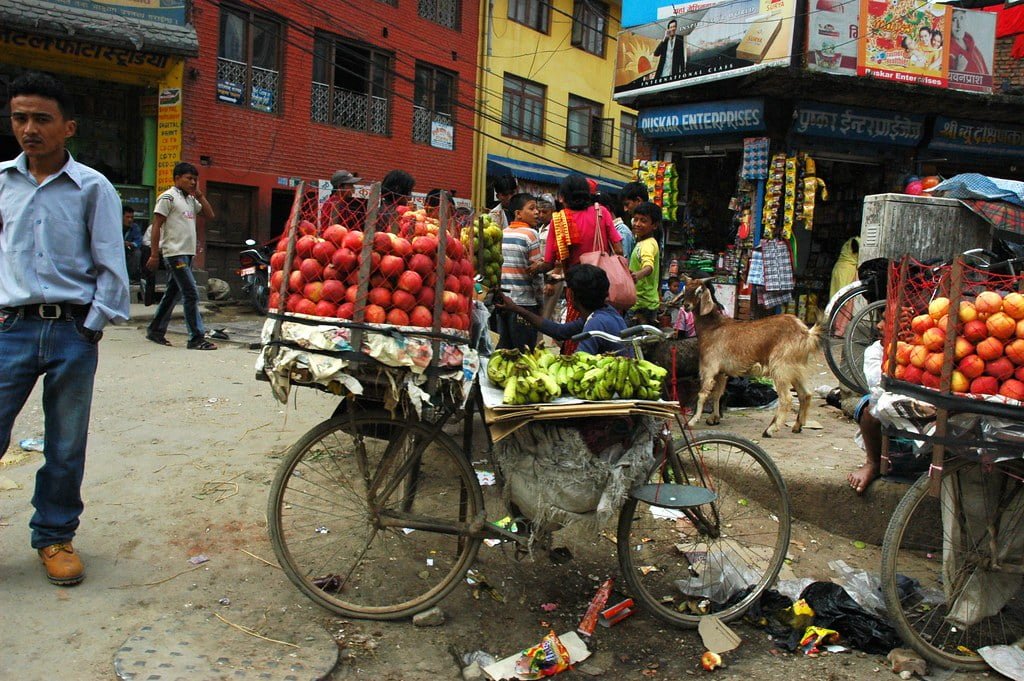 A bustling street market scene with vendors selling fruit from bicycles, a stray goat wandering among scattered litter, and people engaged in commerce in front of colorful storefronts.