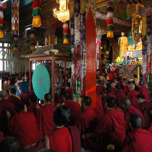 A group of monks in red robes gathers inside a richly decorated Buddhist temple, with some holding large ceremonial drums.