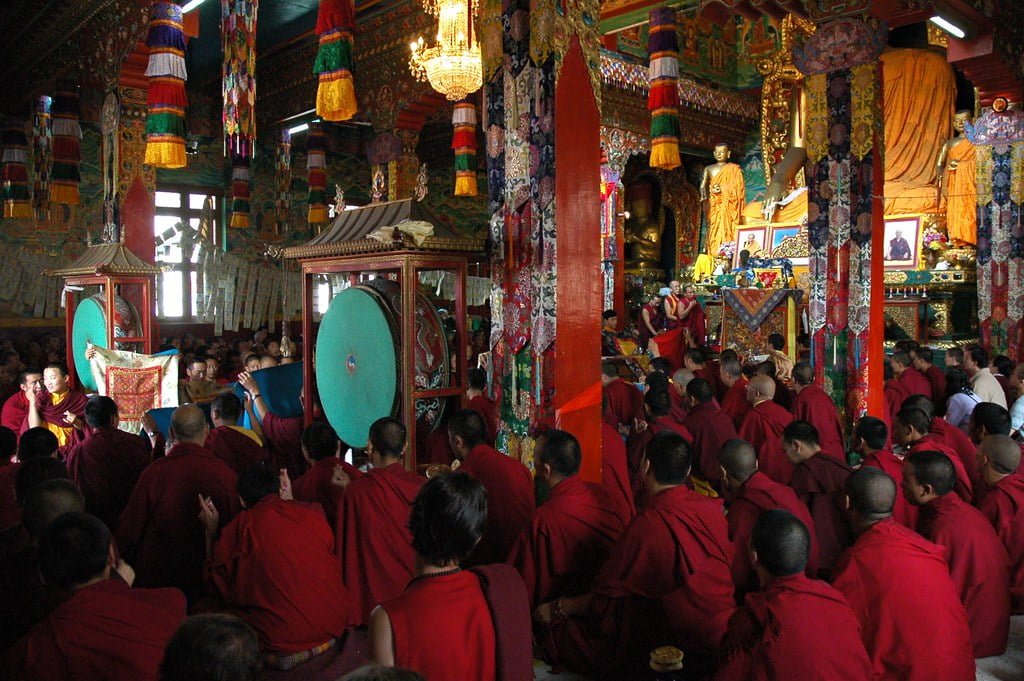A group of monks in red robes gathers inside a richly decorated Buddhist temple, with some holding large ceremonial drums.