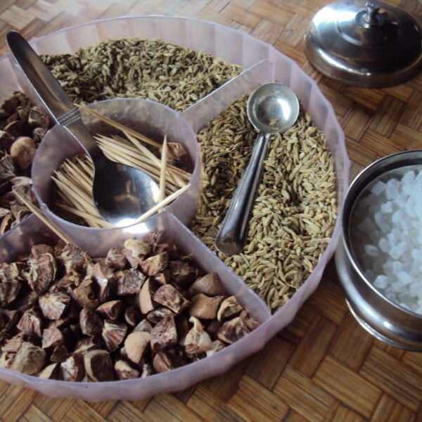 A variety of spices and condiments displayed with spoons on a woven mat, including crushed brown shells, whole cumin seeds, toothpicks, and a metal bowl of white rock salt. A small bell with a push-button mechanism is also visible to the side.