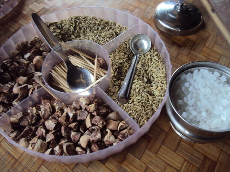 A variety of spices and condiments displayed with spoons on a woven mat, including crushed brown shells, whole cumin seeds, toothpicks, and a metal bowl of white rock salt. A small bell with a push-button mechanism is also visible to the side.