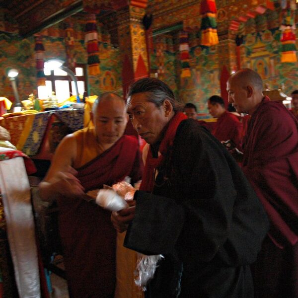 Alt text: A group of monks in traditional maroon robes engaged in a ceremony inside a vibrant, colorfully decorated temple. One monk is seated at a table while others stand or move about, some appearing to be involved in a ritualistic activity.