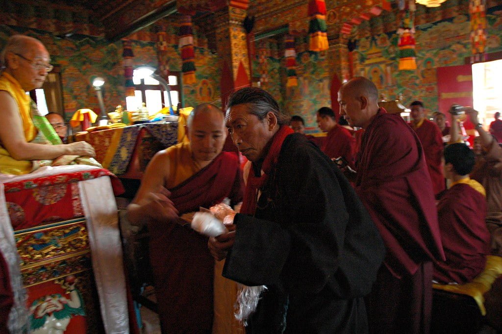Alt text: A group of monks in traditional maroon robes engaged in a ceremony inside a vibrant, colorfully decorated temple. One monk is seated at a table while others stand or move about, some appearing to be involved in a ritualistic activity.