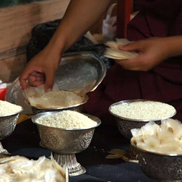 A person in a deep maroon robe sorting and handling what appears to be grains or offerings in metallic bowls on a table, implying a ritualistic or cultural activity.