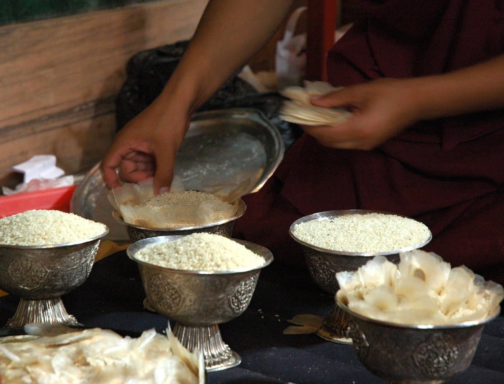 A person in a deep maroon robe sorting and handling what appears to be grains or offerings in metallic bowls on a table, implying a ritualistic or cultural activity.
