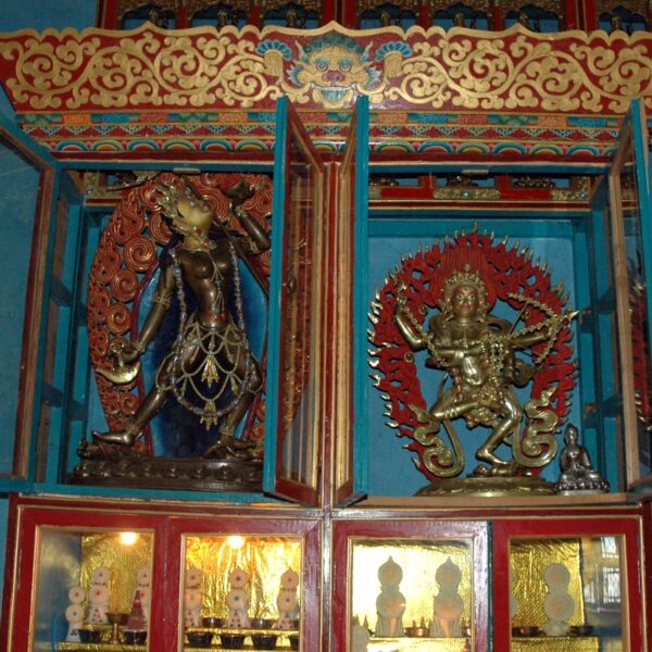 Two ornate, colorful statues of Hindu deities in a shrine with intricate designs and decorations. The statue on the left is dark blue with gold accents, surrounded by a fiery halo, and the one on the right is silver-toned with multiple arms, also surrounded by a fiery halo. Decorative motifs and smaller figures are visible in the background.