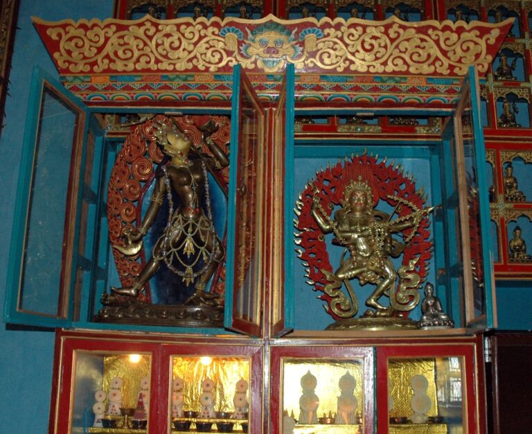 Two ornate, colorful statues of Hindu deities in a shrine with intricate designs and decorations. The statue on the left is dark blue with gold accents, surrounded by a fiery halo, and the one on the right is silver-toned with multiple arms, also surrounded by a fiery halo. Decorative motifs and smaller figures are visible in the background.