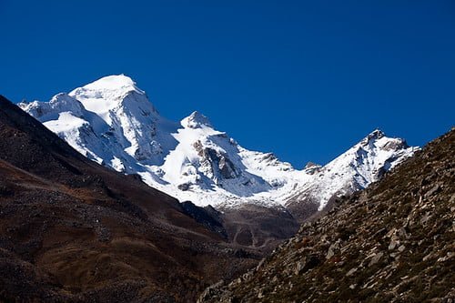Snow-covered mountain peaks under a clear blue sky, with slopes covered in brown vegetation in the foreground.