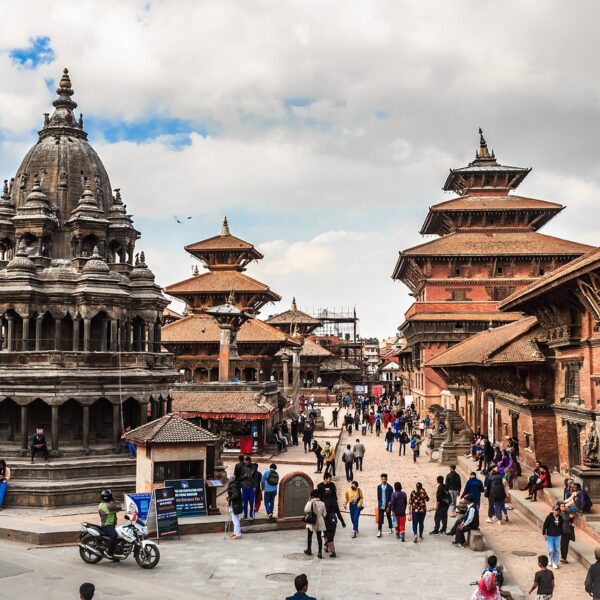An ancient square with intricate multi-tiered temples and historical buildings in Nepal, bustling with locals and tourists against a partly cloudy sky.
