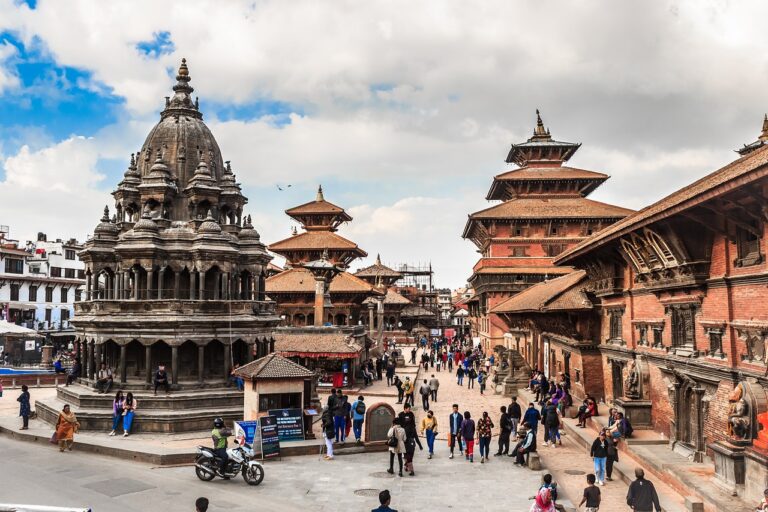An ancient square with intricate multi-tiered temples and historical buildings in Nepal, bustling with locals and tourists against a partly cloudy sky.