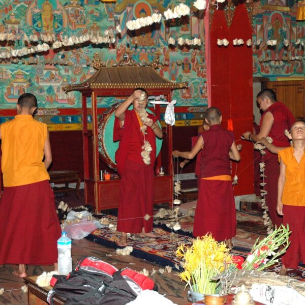 Young Buddhist monks in traditional robes engaged in decoration or ritual preparation inside a colorful monastery with Tibetan mural art on the walls.